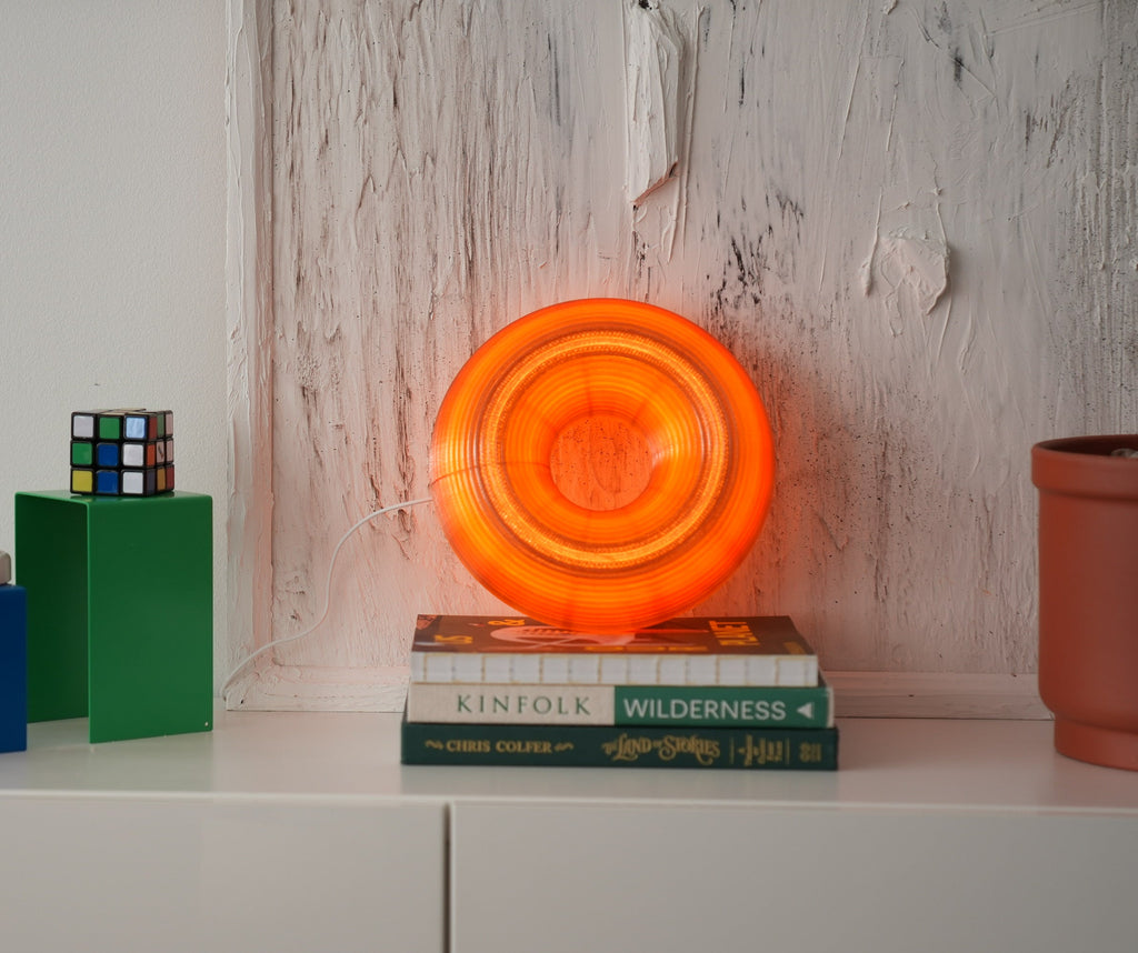 An orange glowing ring-shaped light or lamp sits on top of two books on a white shelf, styled with colorful blocks, a small toy, and a potted plant against a textured white wall.
